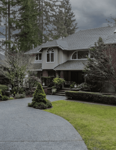 A large brick house with multiple peaks and a separate garage, surrounded by manicured gardens and tall trees under an overcast sky.