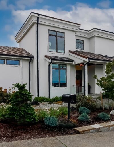 Modern two-story white house with a flat roof, landscaped garden, stone pathway, and outdoor seating area under a clear blue sky.