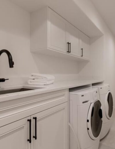 A modern laundry room with white cabinets, a black faucet, a sink, folded towels on the countertop, and a washer and dryer set against a beige-tiled floor. An open door leads to another room.