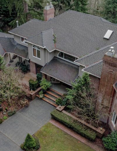 Aerial view of a large suburban home with a gray roof, multiple sections, a brick chimney, and surrounded by trees.