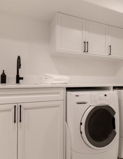 A minimalist laundry room with white cabinets, a black faucet, and a countertop. Below the counter are a front-loading washing machine and dryer set, with folded towels placed above.