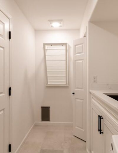 A narrow, well-lit laundry room with white walls, a black faucet on a white sink, a closed white door, wall-mounted cabinet, and a small window with blinds.