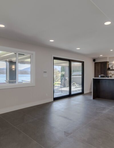 Modern, spacious living room with large windows overlooking a lake, featuring gray tile floors and a dark kitchen island.