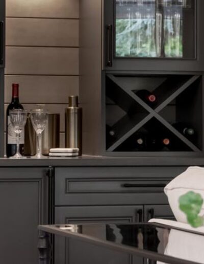 A modern home bar with dark cabinets, wine bottles stored in a crisscross rack, glasses, and a cocktail shaker on the counter, with a potted plant in the foreground.