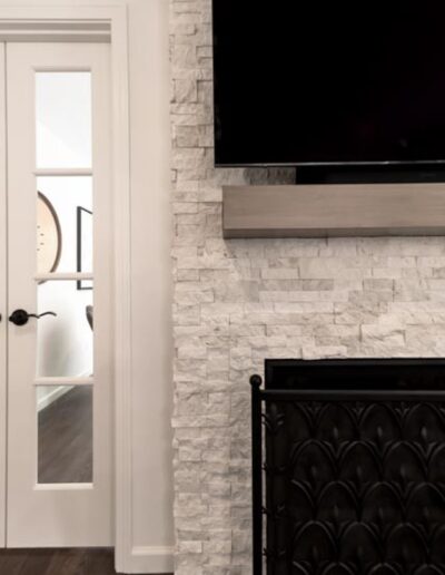 A wall-mounted flat-screen TV above a textured white stone fireplace with a black iron screen. A glass-paneled door is on the left, leading to another room with a visible frame in the background.