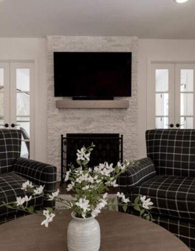 Two checkered armchairs are placed in front of a coffee table with a vase of flowers in a minimalist living room. A TV is mounted above a fireplace between two French doors.