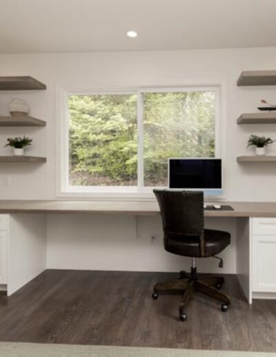 A home office with a large desk beneath a window, surrounded by floating shelves. A computer monitor, an office chair, and potted plants are on the desk. The walls and cabinetry are white.