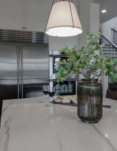 Modern kitchen interior with stainless steel refrigerator, marble countertop, hanging lamp, green potted plant, and two wine glasses.