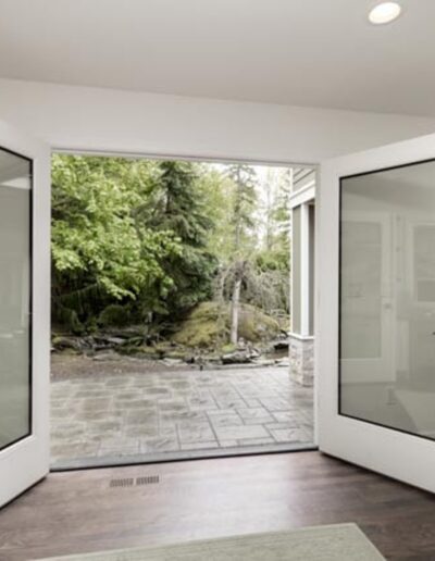 Open double glass doors lead from a modern interior to a stone patio outside, surrounded by lush greenery.