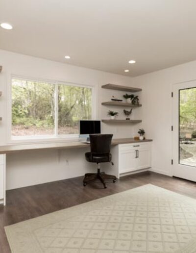 A modern home office with a large window, built-in desk, shelving, and a black office chair. French doors lead outside. White cabinets and light wood accents complement the space.