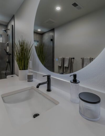 Modern bathroom interior with a white sink, black faucet, and a large mirror, complemented by decorative green plants and grey towels.