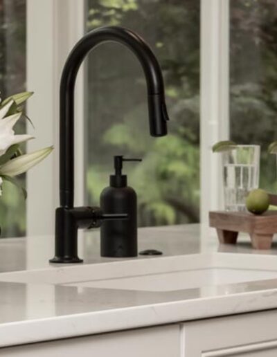 A modern kitchen sink with a black faucet is displayed, featuring a vase of white flowers on the counter and a wooden tray with glasses of water and a pitcher containing lime slices.