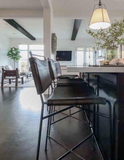 Modern open-plan kitchen and living room with bar stools, dark countertops, a chandelier, and a view into a well-furnished living area.