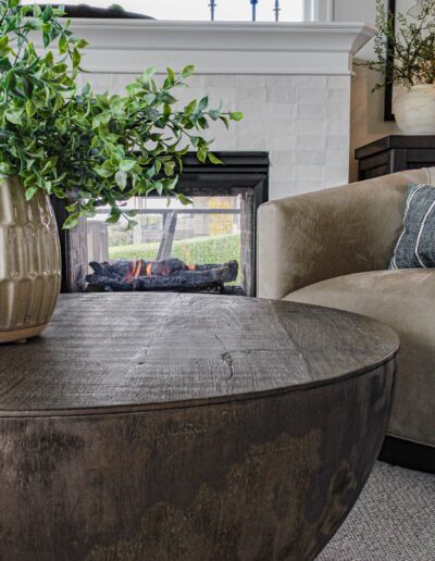 A cozy living room corner with a modern fireplace, a round wooden coffee table, and a beige armchair, accented by a plush green plant on a nearby shelf.