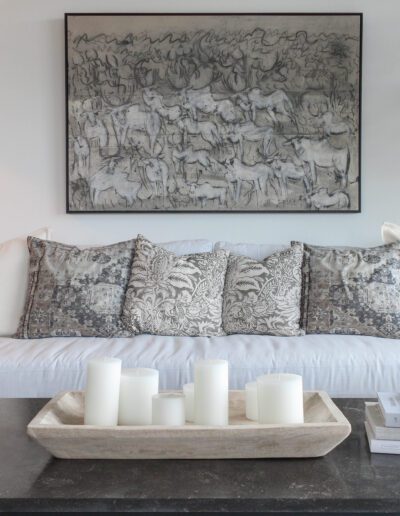 A modern living room featuring a white sofa with decorative pillows, a coffee table with candles and books, and a large abstract art piece on the wall.