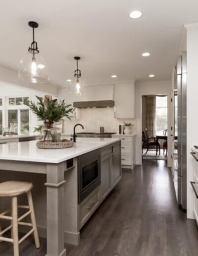 A modern kitchen with white cabinets, a large island with two wooden stools, pendant lights, and stainless steel appliances. A window on the left side provides natural light.