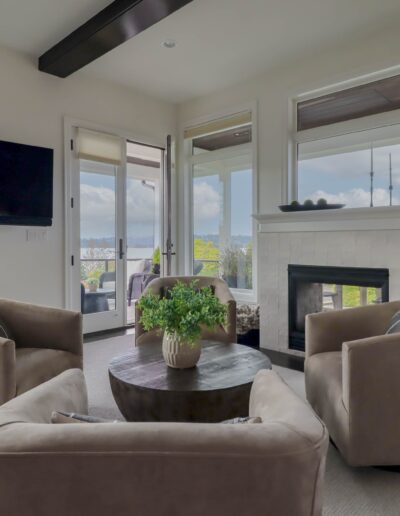 Modern living room with beige armchairs, a round wooden coffee table, a fireplace, and large windows offering a view of the outside.