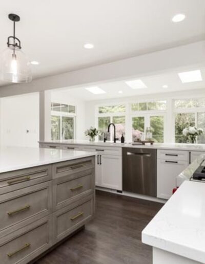 Modern kitchen with a large island featuring a sink and storage, pendant lights, white countertops, stainless steel appliances, and large windows providing natural light.