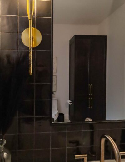 A modern bathroom featuring dark tiles, a black cabinet, brass fixtures, and a decorative feather arrangement on the left.