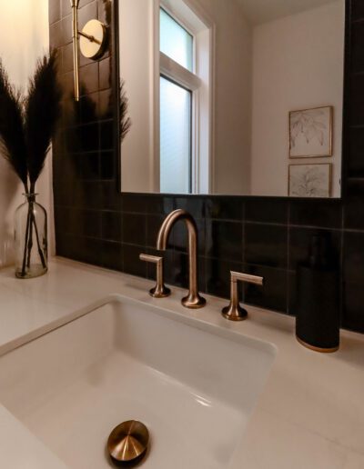 A modern bathroom sink with brass faucets, surrounded by black tiles, accompanied by decorative items and artwork.