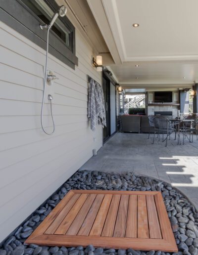 An outdoor shower with a wooden floor mat surrounded by grey stones next to a sliding glass door leading to a modern home patio.