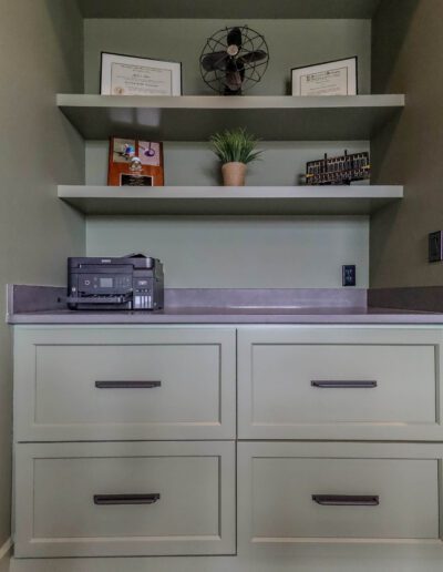 A modern office nook featuring a white built-in desk with drawers, floating shelves above holding a printer, decorative items, and plants, all against a gray wall.