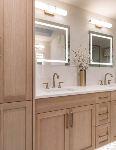 Modern bathroom with light wood cabinets, double vanity sinks, gold accents, and a lighted mirror. decor includes a vase of dried flowers.