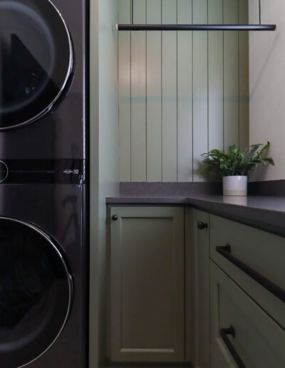 A modern laundry room featuring a stacked washer and dryer to the left and built-in green cabinets with a countertop and a small potted plant to the right.