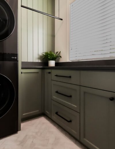 Modern laundry room with stacked black washing machine and dryer, dark green cabinets, and a sink by a window with blinds.