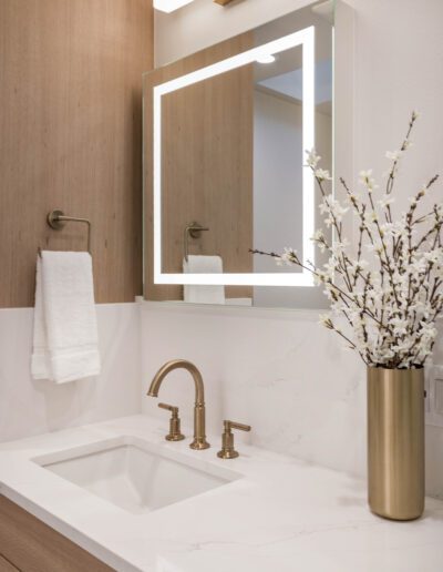 Modern bathroom interior with a backlit mirror, white marble countertop, gold faucet, and a vase of white flowers.