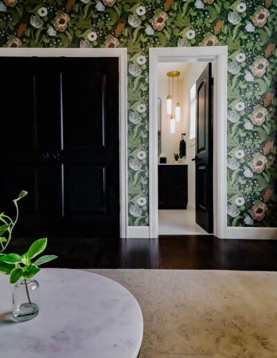 Interior of a room with floral wallpaper, featuring a white marble table with two glass vases and green plants, a green armchair, and dark doors.