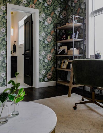 Elegant home office with floral wallpaper, a wooden shelf stocked with books, a marble-topped table, and a green upholstered chair near a window.