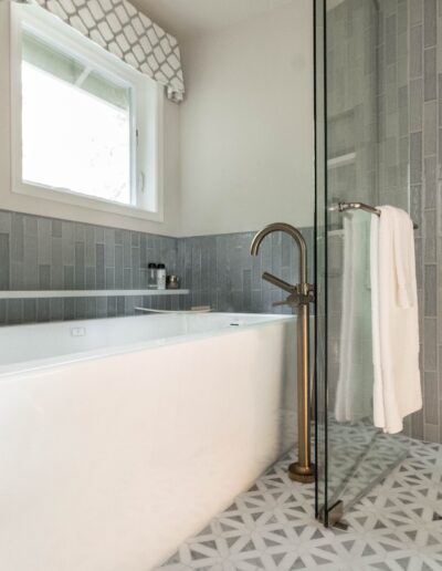 A modern bathroom featuring a standalone bathtub with a glass shower partition, patterned floor tiles, and a small window.