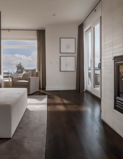 Modern bedroom with a white bed, dark hardwood floor, and a double-sided fireplace, opening to a sunny balcony with a view of the sea.