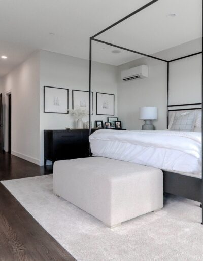 Modern bedroom with a large bed framed by a black canopy, white bedding, dark wood furniture, a gray bench at the foot of the bed, and a potted plant on the left.