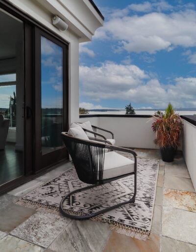 Modern balcony with a stylish black rocking chair on a patterned rug, stone flooring, potted plants, and a clear sky in the background.