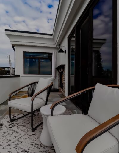 Modern patio area with two stylish chairs, a rug, and a firewood storage next to a glass door, under a cloudy sky.