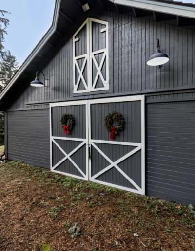 A grey barn with white cross-hatch door designs and festive wreaths, flanked by white lamps, against a woodland backdrop.