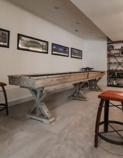 Interior of a game room featuring a shuffleboard table, three bar stools, framed photos on the wall, and a shelving unit with various items.