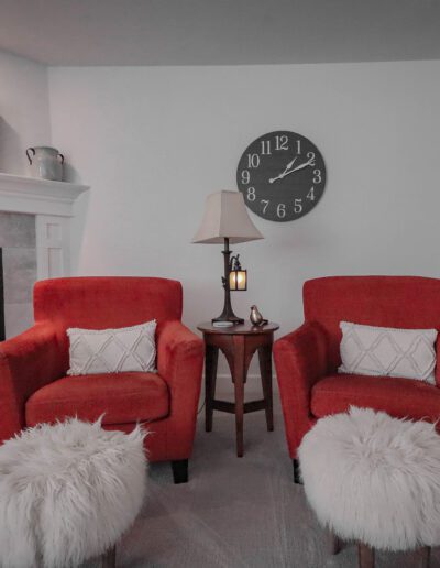 Two red armchairs with white cushions in front of a fireplace, flanked by furry white stools and a small table with a lamp, under a large clock on a gray wall.