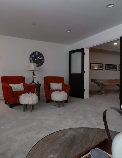 A cozy living room featuring two orange chairs by a fireplace, a large tv on the wall, and a decorative clock above a fireplace mantel adorned with photos.