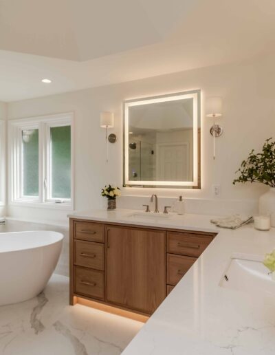 Luxurious bathroom with a freestanding bathtub, large vanity with wood cabinets, backlit mirror, and green plants. Marble floor and countertops complement the elegant decor.