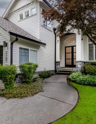 Modern two-story white house with a dark garage door, surrounded by manicured shrubs and a lush green lawn. The entrance features a tall glass-paneled door and arched window.