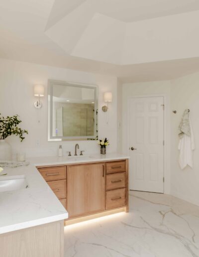 A modern bathroom with a white marble countertop, wooden cabinets, a large mirror, wall sconces, and decorative plants.