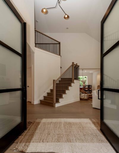 A modern entryway with open double glass doors, a woven rug, and a staircase leading to an upper level. A living area with seating is visible in the background.