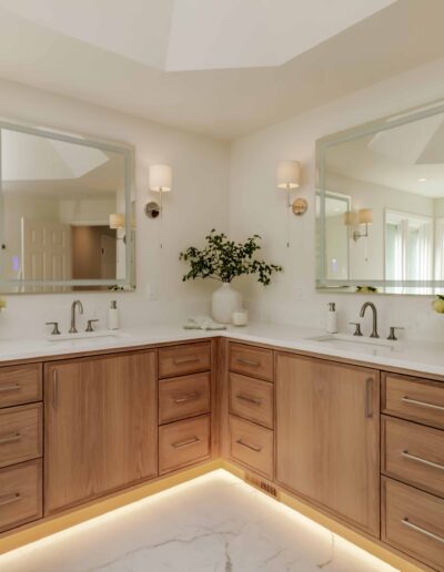 A bathroom with a corner double vanity featuring two mirrors, wooden cabinets, under-cabinet lighting, and decorative plants on the countertops.