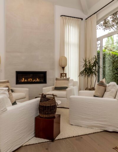 A cozy living room with four white armchairs around a modern fireplace. Large window with draped curtains on the right. Decor includes plants and a basket on a small table.