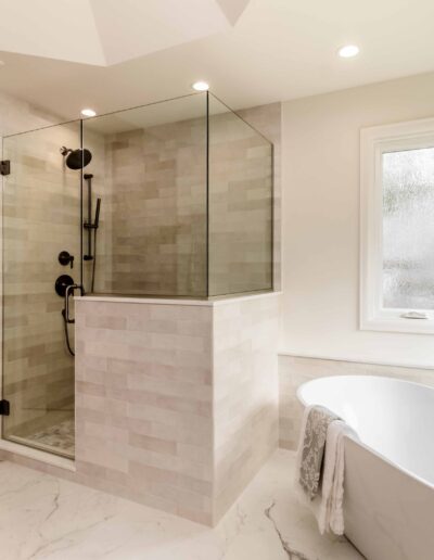 A modern bathroom with a glass-enclosed shower, freestanding bathtub, and large windows. The space features light-colored tiles and marble flooring.