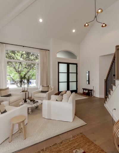 A modern living room with white furniture, a large window, wooden flooring, and a staircase. A modern light fixture hangs from the ceiling.