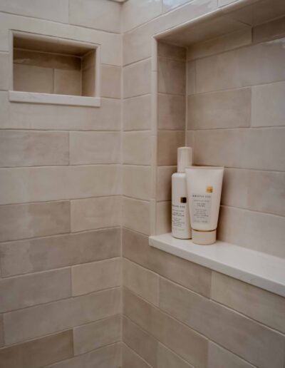 A corner of a tiled shower with two built-in shelves. The lower shelf holds three bottles of white personal care products.